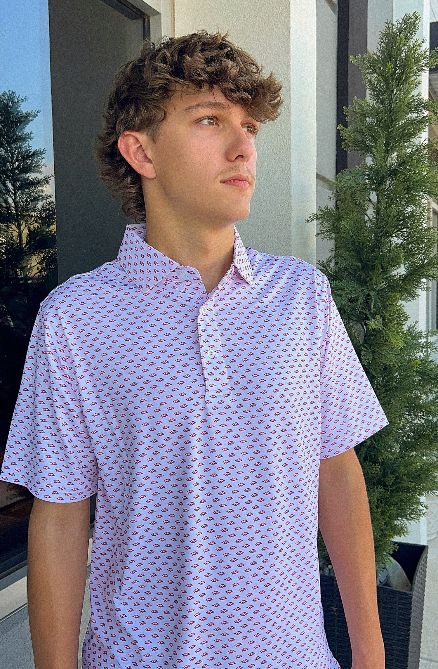 Young man wearing a patterned shirt standing outdoors with a building and tree in the background