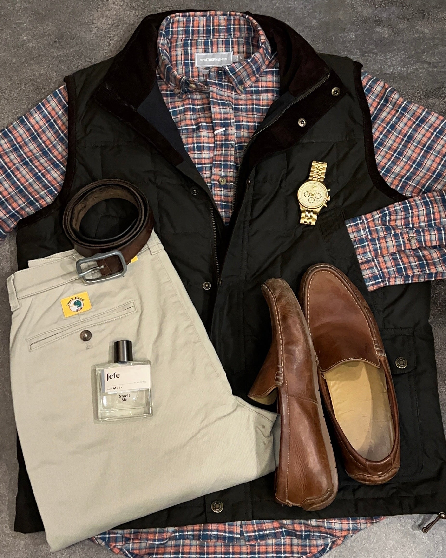 Collection of clothing and accessories including a plaid shirt, vest, pants, loafers, and a belt on a gray surface.