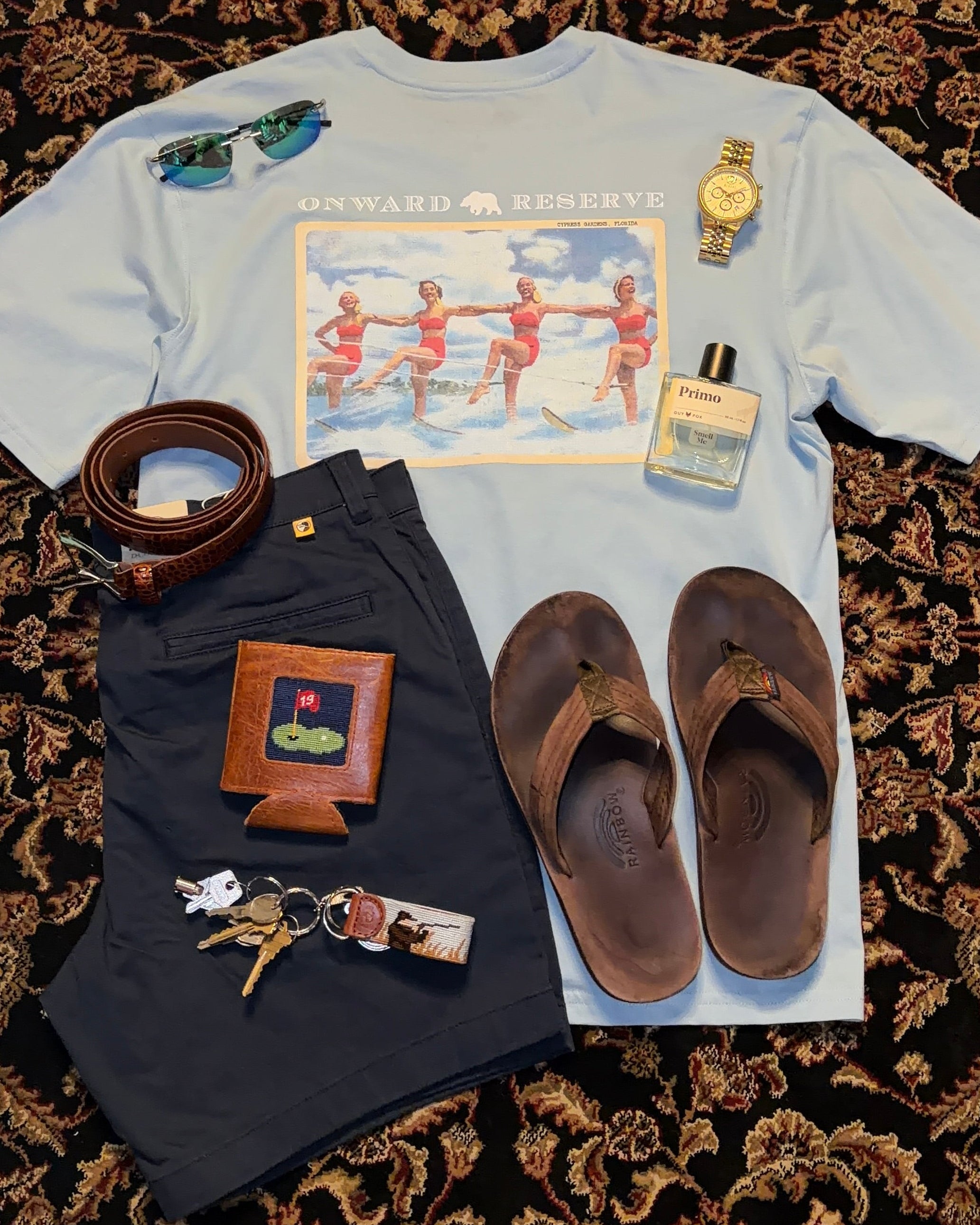 Collection of items including a shirt with a graphic, sandals, a belt, wallet, keys, and a watch on a patterned surface.