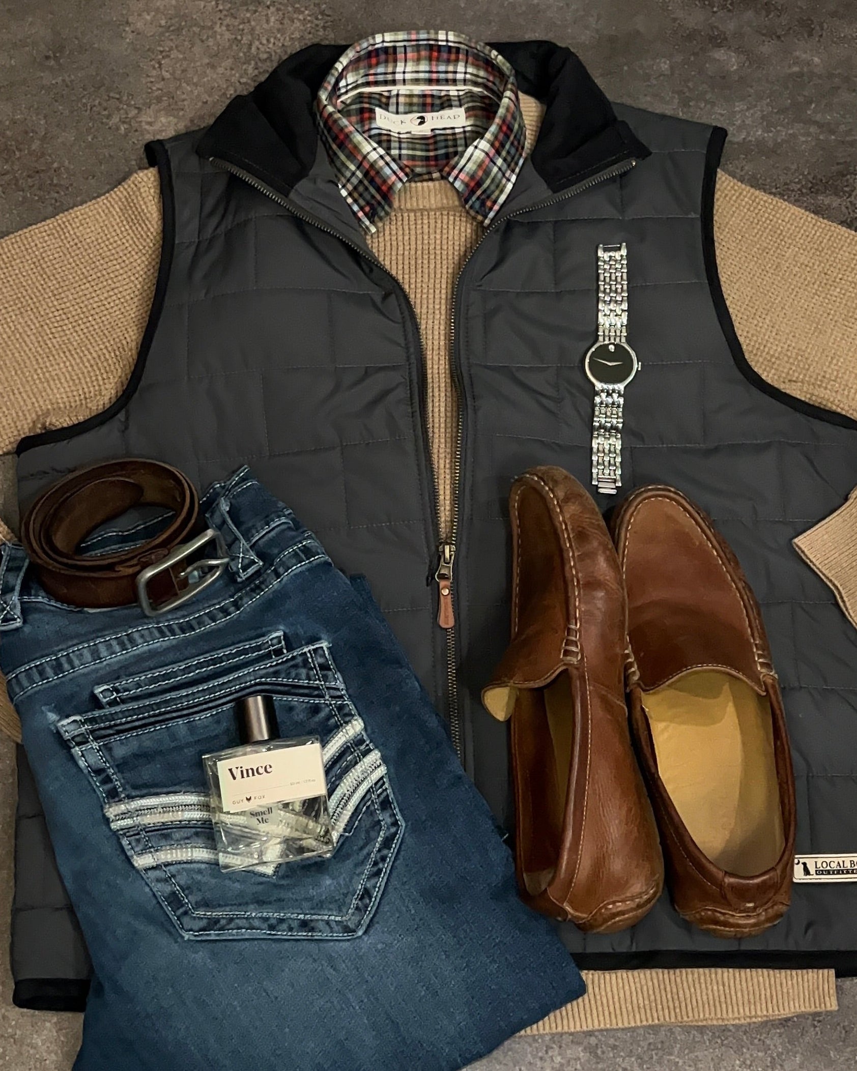 Collection of men's clothing and accessories including a vest, jeans, shoes, and belt on a gray surface.