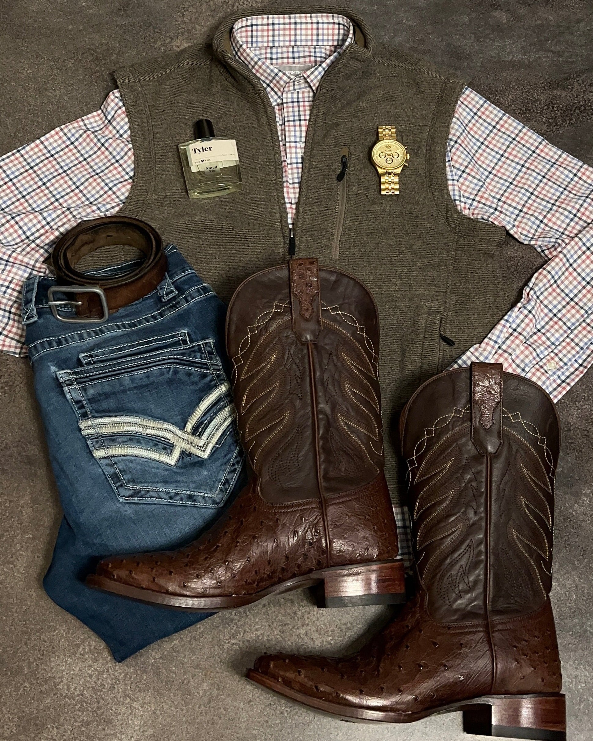 Cowboy boots, jeans, vest, and shirt laid out on a concrete floor.