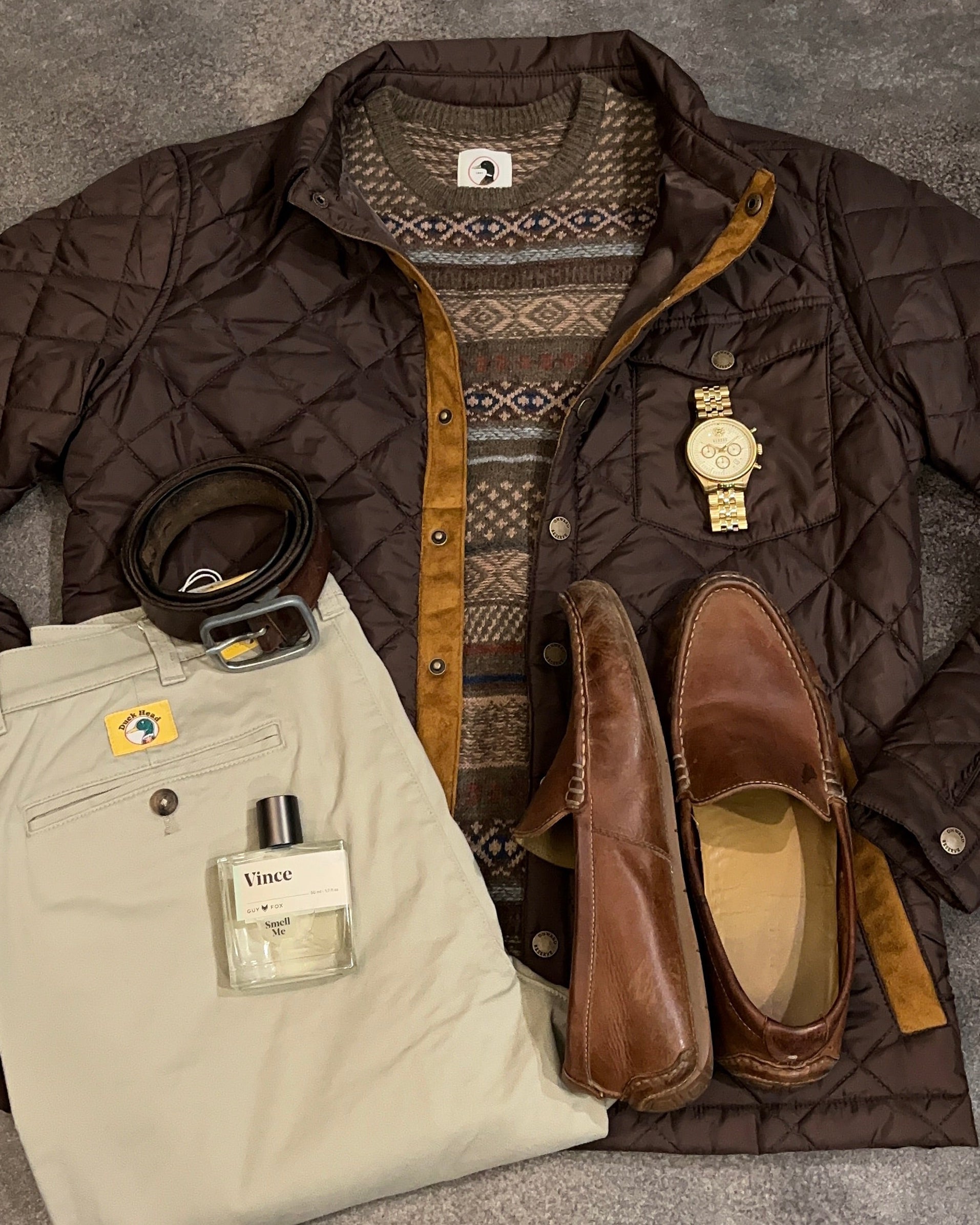 Fall fashion items including a quilted jacket, patterned sweater, pants, loafers, and a belt on a gray surface.