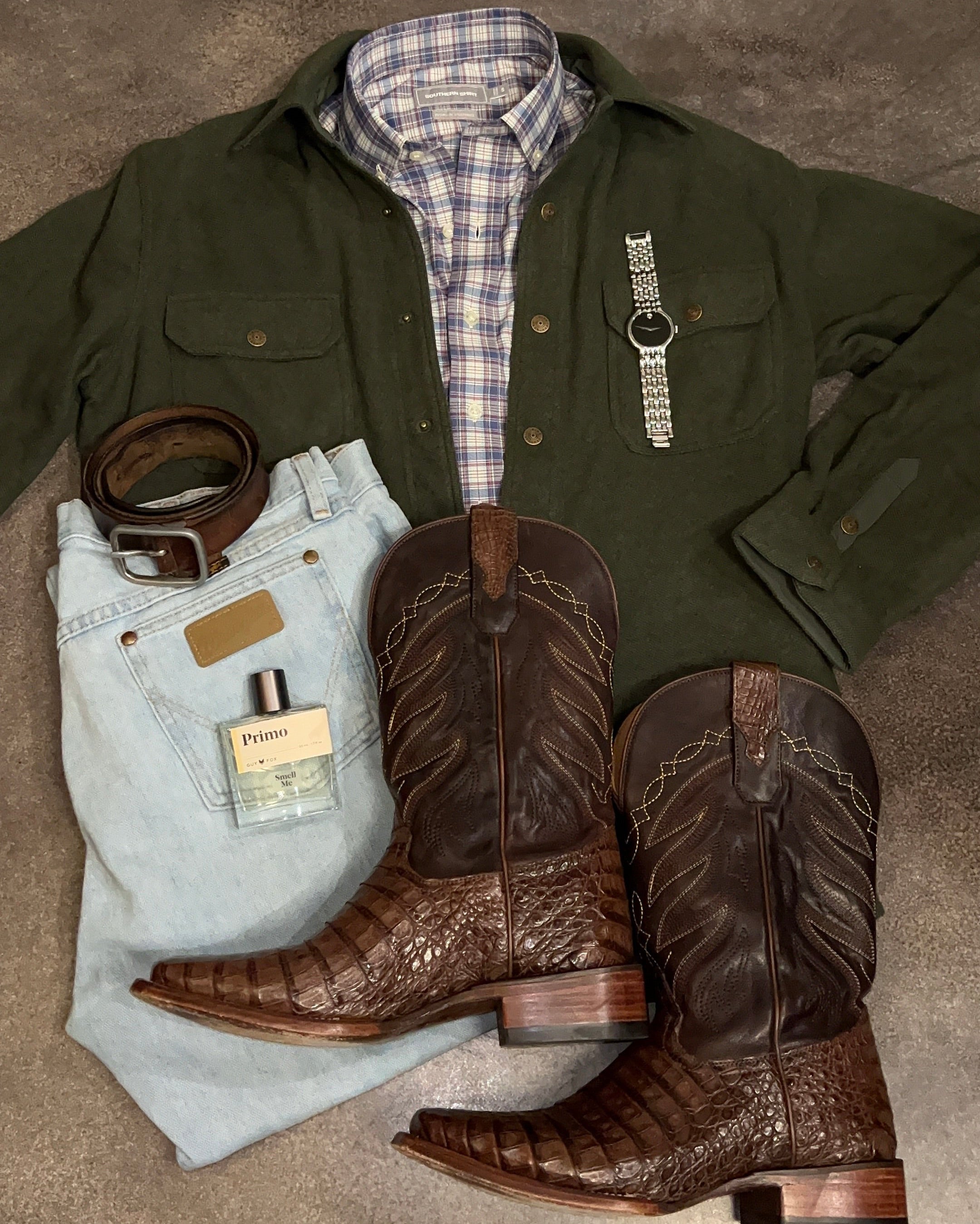 Green jacket, plaid shirt, jeans, brown cowboy boots, belt, and perfume bottle on a gray surface.
