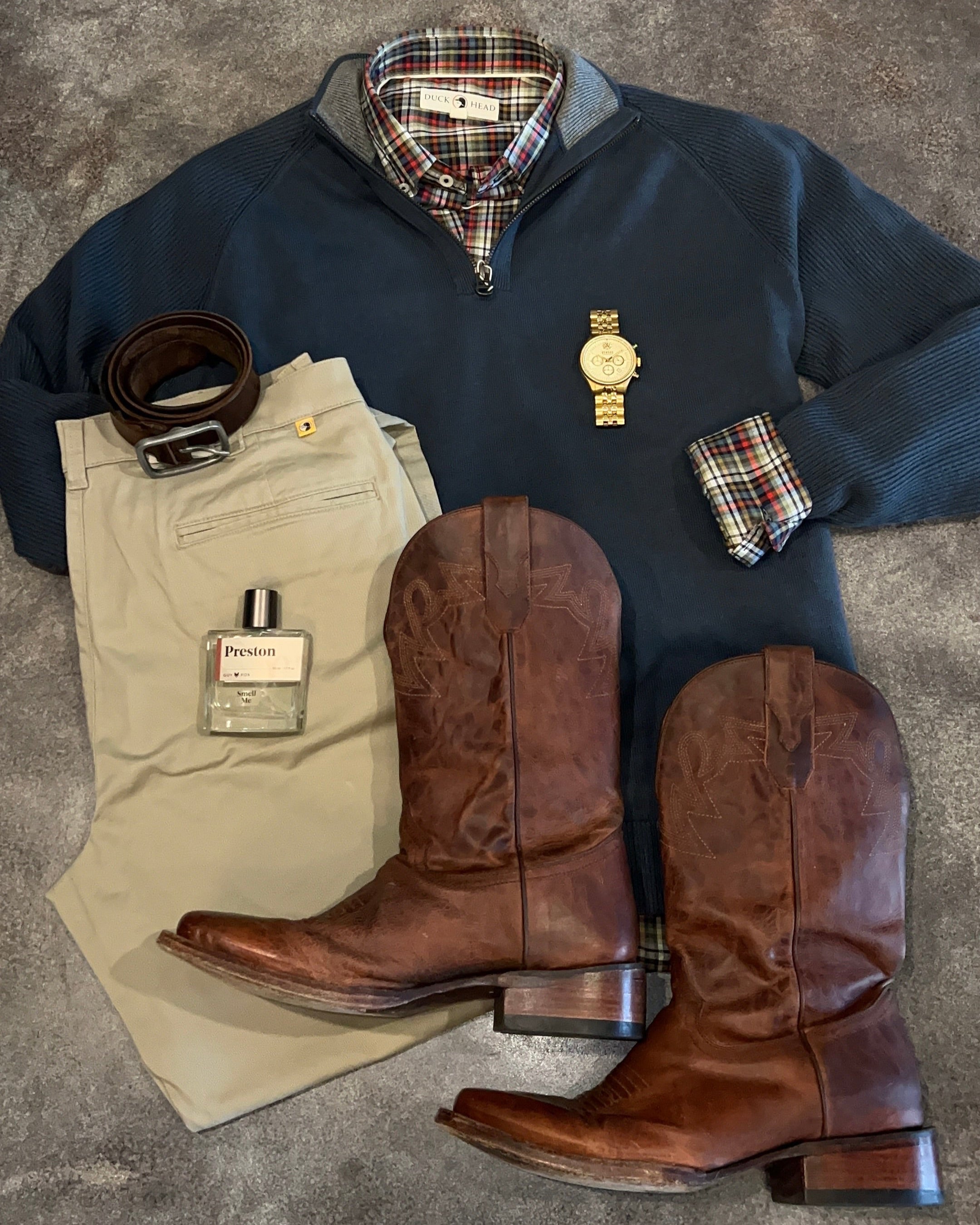 Collection of men's clothing and accessories including a sweater, pants, boots, belt, and perfume bottle on a textured surface.