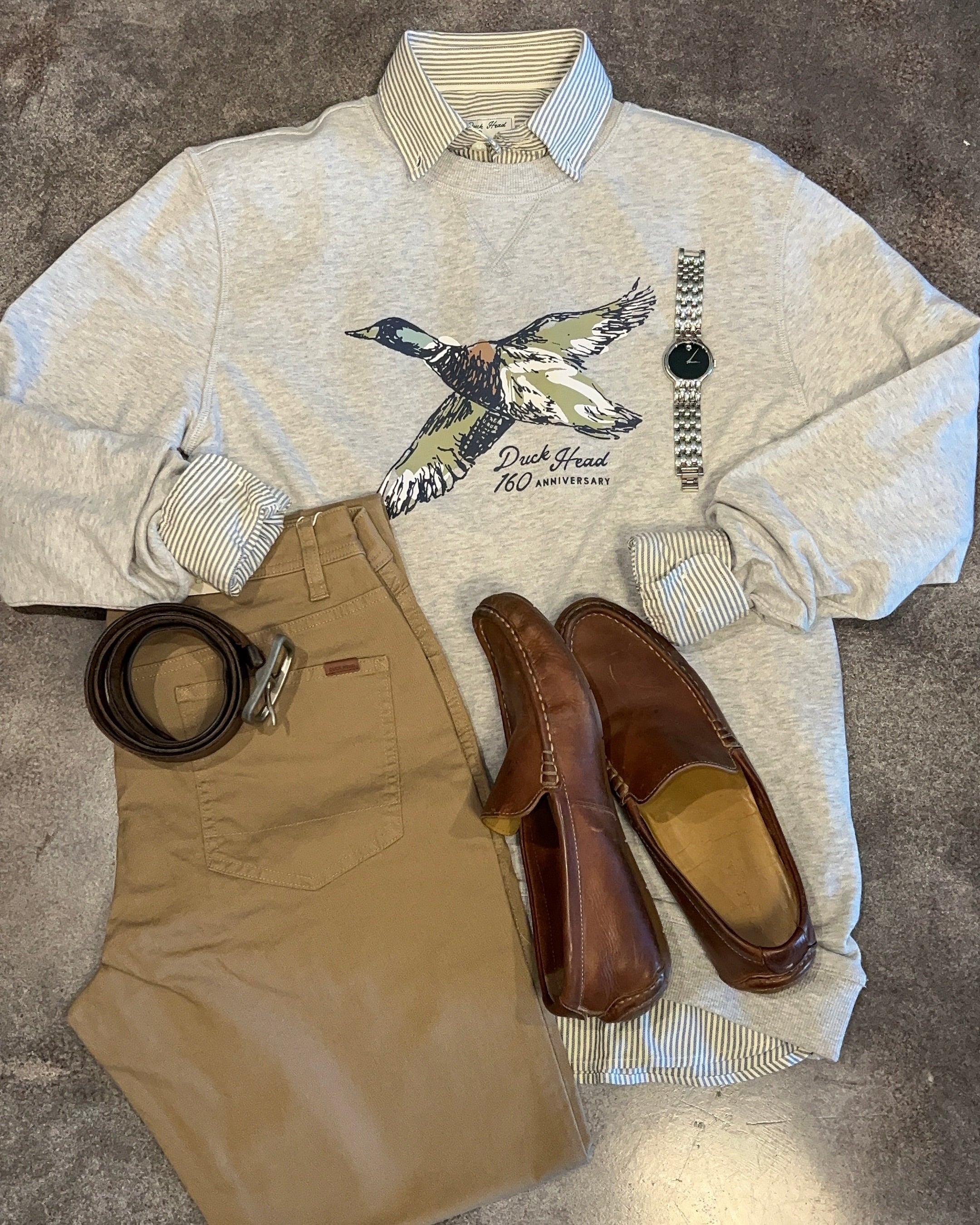 Collage of a shirt with a graphic design, brown pants, shoes, and a belt on a gray surface.