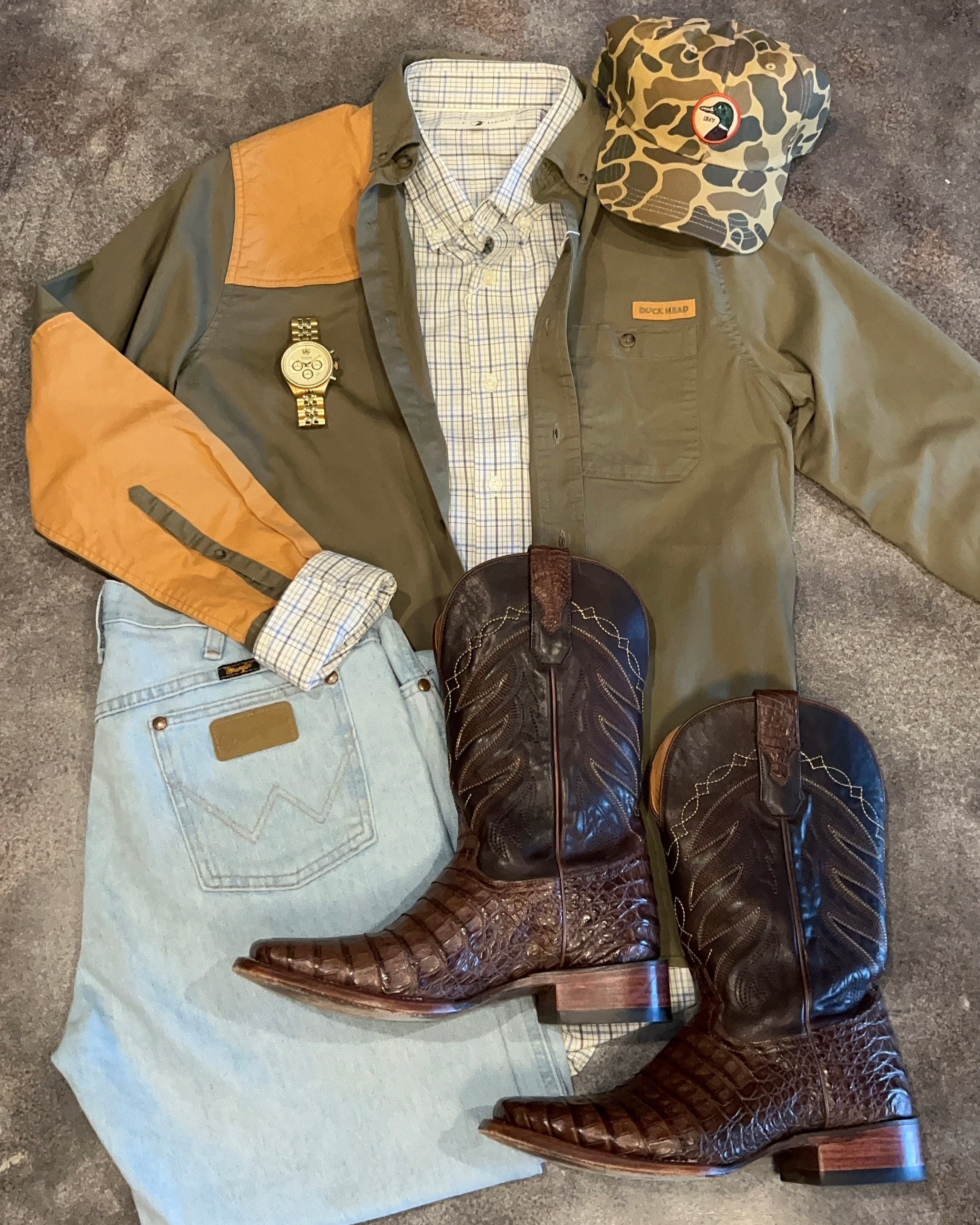 Collection of cowboy boots, jacket, shirt, jeans, and watch on a concrete floor.