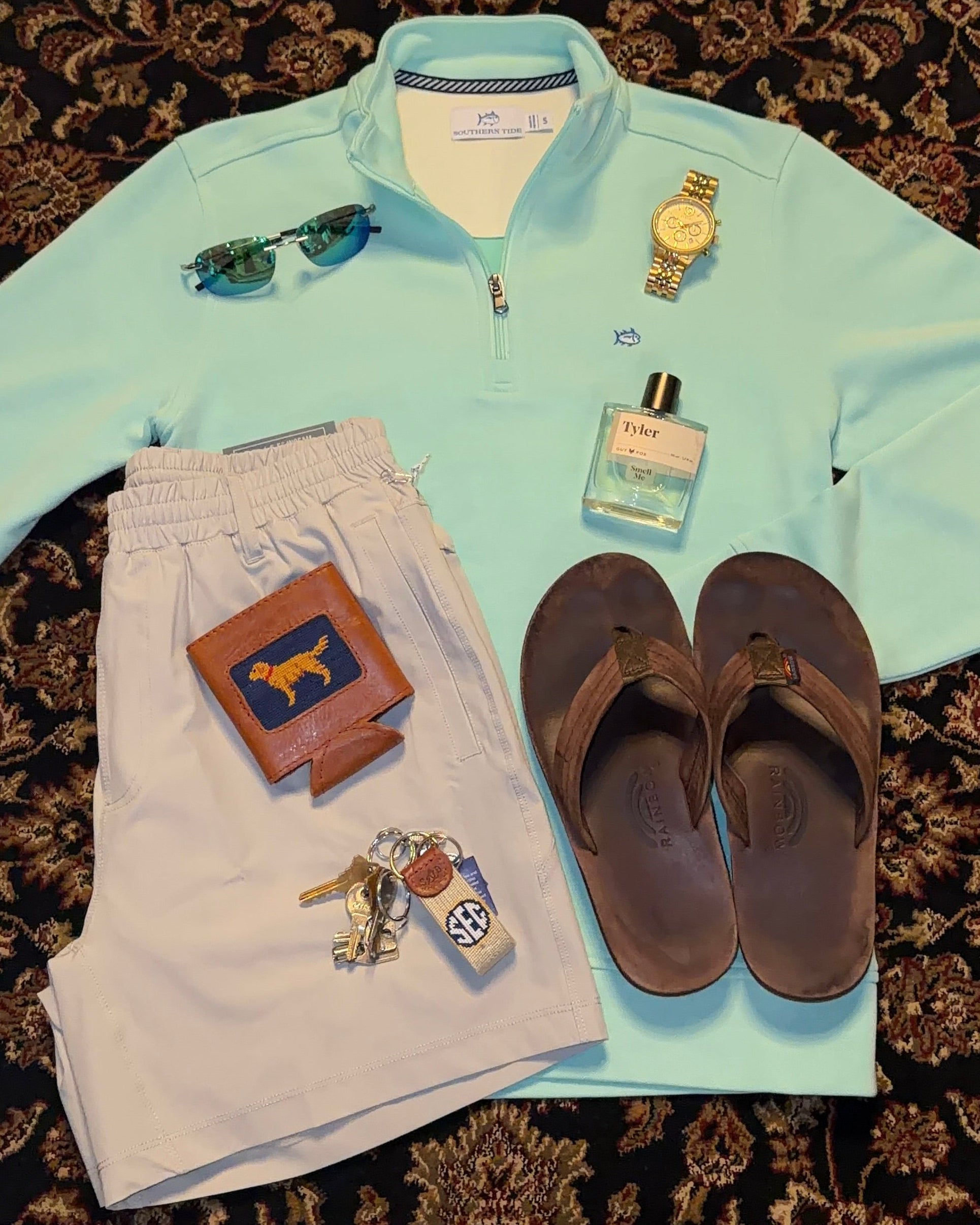Light blue shirt, white pants, sandals, wallet, keys, and perfume bottle on a patterned rug.